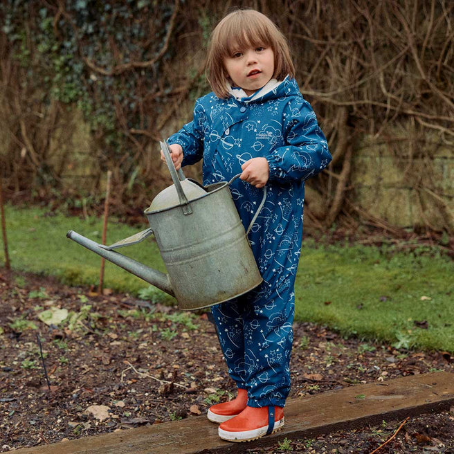 A child in a navy 3 in 1 Waterproof Scampsuit, patterned with planets, stars & clouds, stands on wood holding a watering can, in a garden.